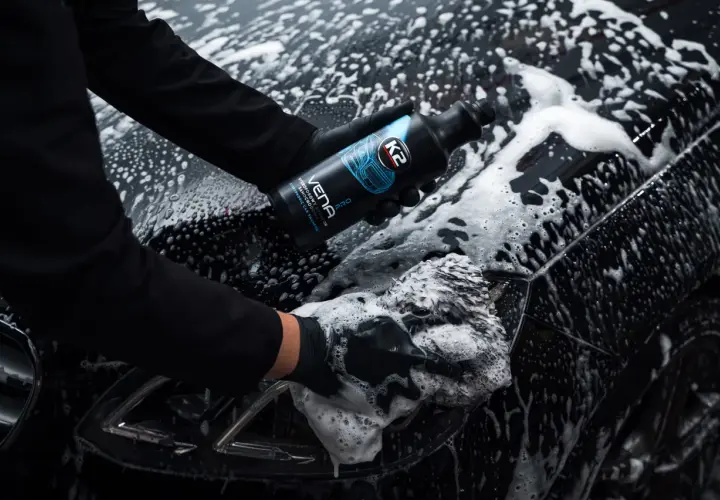 Ręce w czarnych rękawiczkach myją czarne auto pianą, trzymając butelkę szamponu.