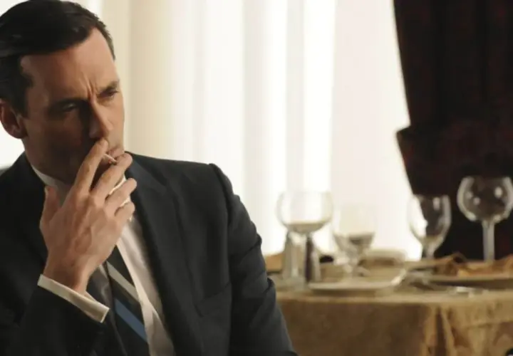 Jon Hamm, znany z filmów, seriali i programów, w eleganckim garniturze pali papierosa przy stole.