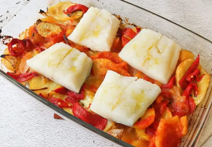 Cuatro porciones de bacalao al horno reposan sobre patatas y pimientos asados en una fuente de vidrio.