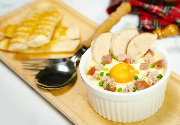 Un œuf cocotte bain-marie, garni de saucisse et de viande hachée, servi avec des bâtonnets de pain grillé.