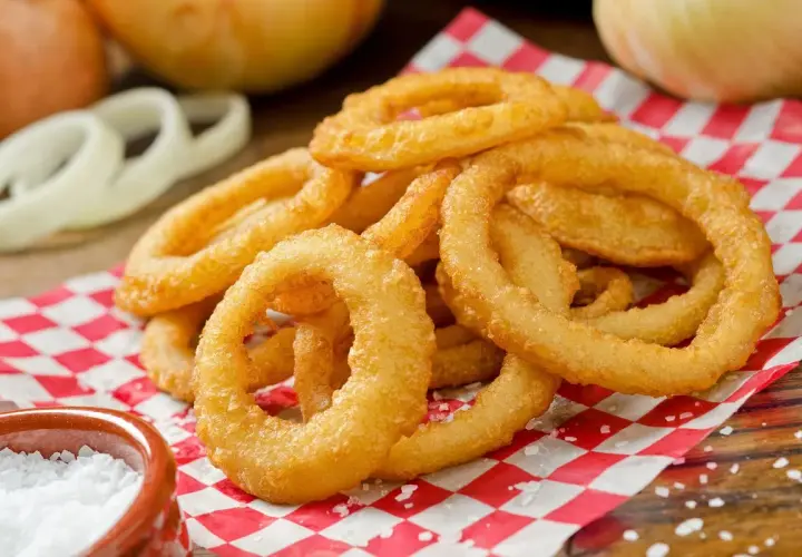 Anelli di cipolla dorati e croccanti, preparati con pastella con acqua frizzante, serviti su carta a quadri rossi e bianchi, con sale grosso a lato.