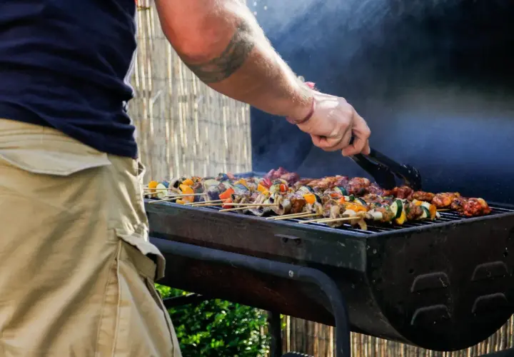 Un homme utilise des pinces pour retourner des brochettes colorées sur un barbecue fumant. C'est le moment idéal pour degraisser le barbecue après ce festin !