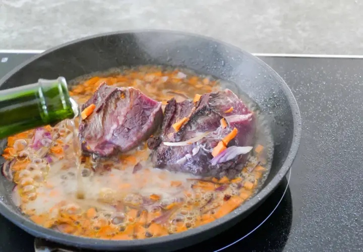 Deux morceaux de joue de bœuf mijotent dans une poêle avec des carottes et des oignons. Un liquide est versé d'une bouteille verte.