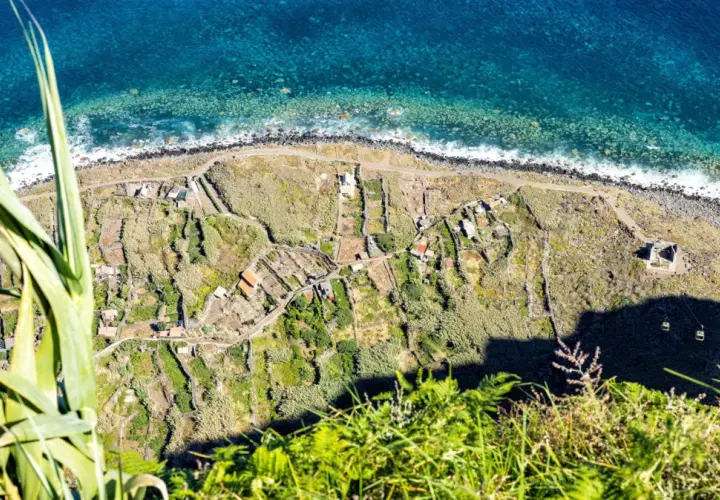 Terraced fields and houses dot the coastline, a glimpse of what to see in Madeira. The turquoise ocean crashes against the rocky shore.