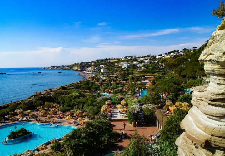 Vista panoramica dell'isola d'Ischia con mare azzurro, piscine, vegetazione lussureggiante e case arroccate sulla collina.