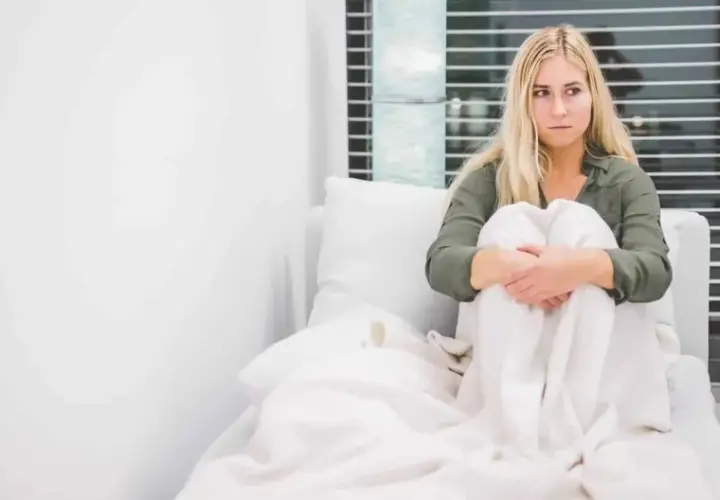 Frau in dissoziativem Stupor, in Decken gehüllt, sitzt auf einem Sofa, Blick leer, Fenster im Hintergrund.