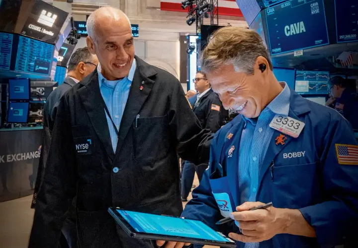Dwóch mężczyzn w kurtkach NYSE śmieje się, patrząc na tablet. Jeden z nich, Bobby, pokazuje coś na ekranie, a drugi, z napisem "skup akcji własnych" na piersi, uśmiecha się szeroko.