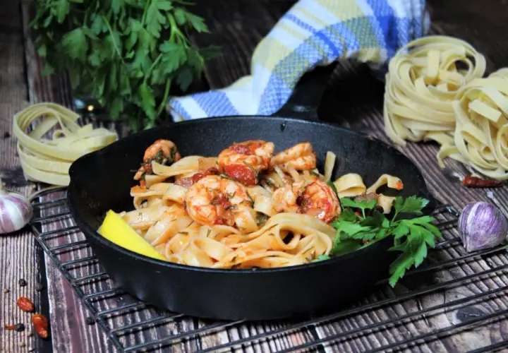 Spaghetti mit Garnelen mediterran in einer Gusseisenpfanne, garniert mit Zitrone und Petersilie. Daneben rohe Pasta, Knoblauch und Chili.