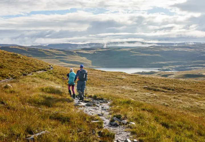 Two hikers trek along the Cape Wrath Trail, a rugged path winding through a vast, misty Scottish landscape with a serene loch in the distance.