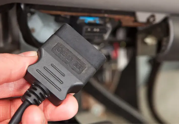 Dłoń trzyma czarny adapter OBD II przed panelem diagnostycznym samochodu.