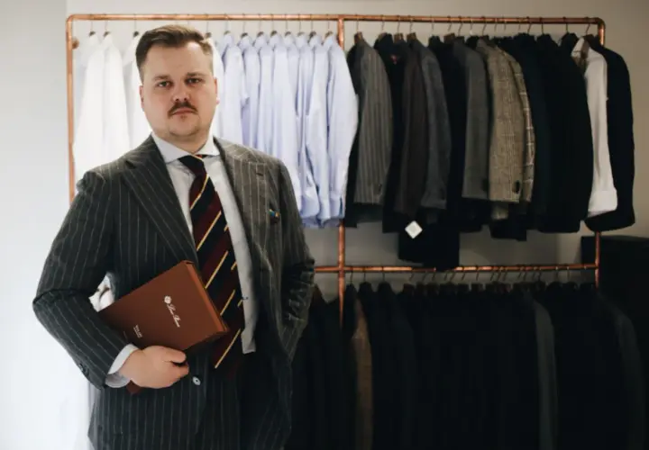 Elegancki mężczyzna w garniturze w prążki trzyma teczkę. Za nim wiszą marynarki i koszule.