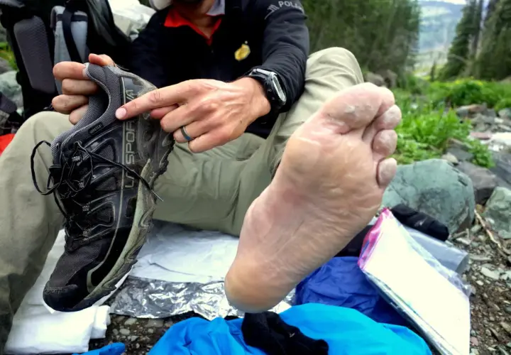 A hiker shows a damp foot next to a muddy shoe, illustrating what causes shoes to dry rot: prolonged exposure to moisture and lack of ventilation.