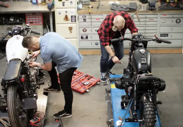 Due uomini lavorano su due moto in un garage. Uno è chino su una moto bianca, l'altro su una nera. Un kit di attrezzi è aperto sul pavimento. Progetto schienalino moto fai da te.