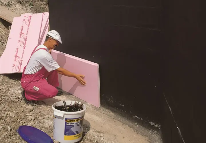 Budowlaniec przykłada różowy styropian do czarnej ściany. Czy styropian przewodzi prąd? Nie, jest izolatorem.