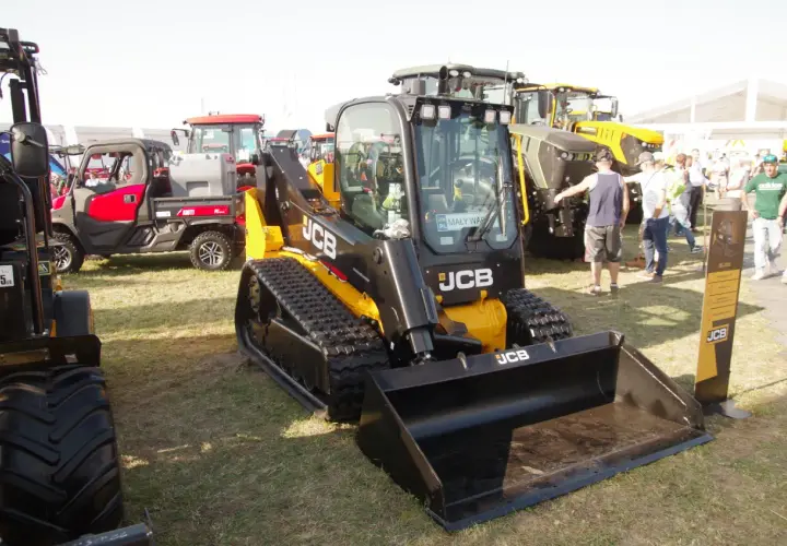 Głównym elementem jest minikoparka gąsienicowa JCB. W tle widać inne maszyny rolnicze i ludzi. Możliwe, że to wystawa, gdzie można sprawdzić jcb polska cennik.