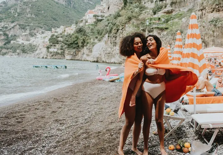 Deux amies rient sur une plage italienne, partageant une serviette orange. Leurs vacances en Italie en famille au bord de la mer sont idylliques.