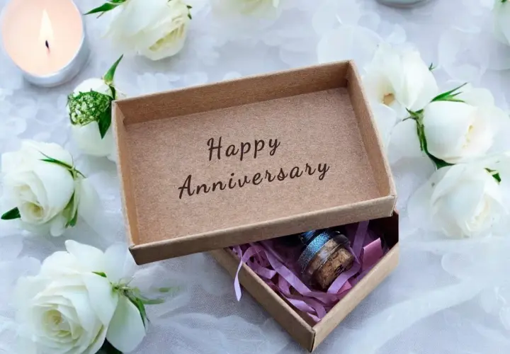 Otwarta szkatułka z napisem "Happy Anniversary" i małą buteleczką w środku, otoczona białymi różami i płonącą świecą. Idealny prezent na 21 rocznicę ślubu.