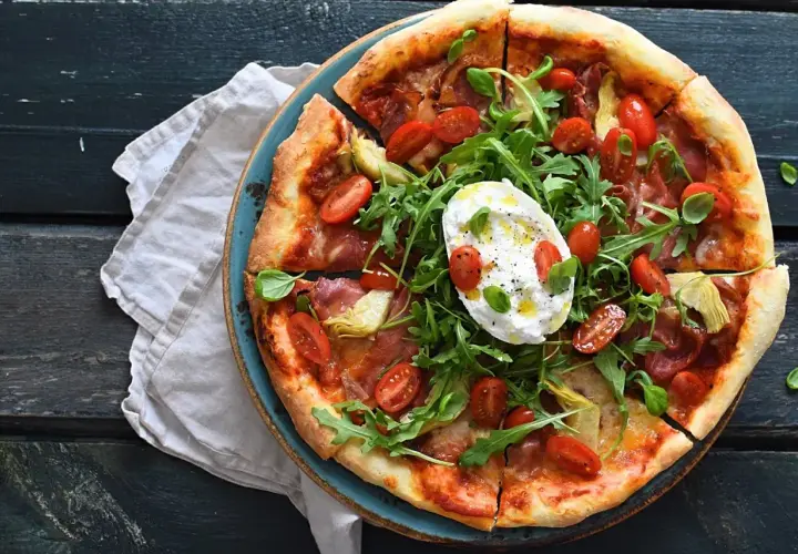 Pizza z mortadelą, rukolą, pomidorkami i burratą na niebieskim talerzu.