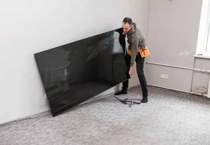 Mężczyzna z pasem narzędziowym przy ścianie trzyma duży, czarny telewizor. Zastanawia się, na jakiej wysokości telewizor będzie najlepiej wyglądał.