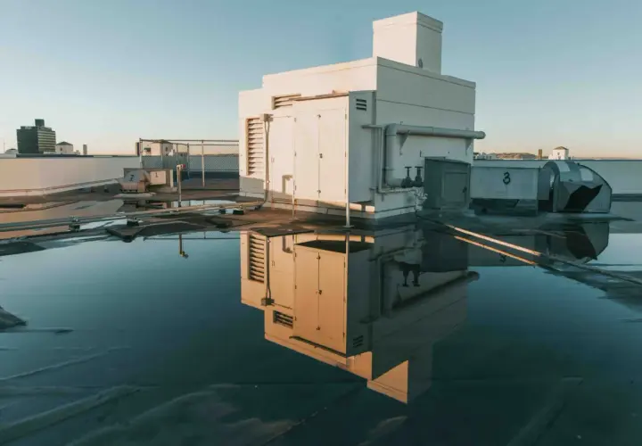 Flachdach mit Wasserpfützen, die ein Gebäude und Lüftungsanlagen spiegeln. Möglicherweise Anzeichen von Flachdach Blasenbildung unter der Oberfläche.
