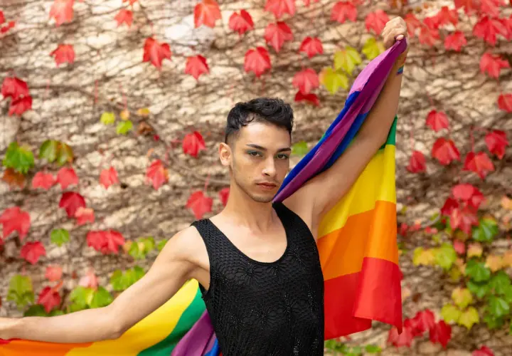 Un giovane con la bandiera arcobaleno, simbolo del **queer significato** di libertà e identità, si staglia contro un muro fiorito.