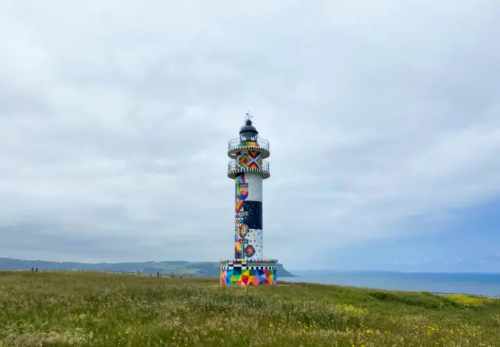 Faro colorido en el cabo de Ajo, con un paisaje verde y el mar al fondo.
