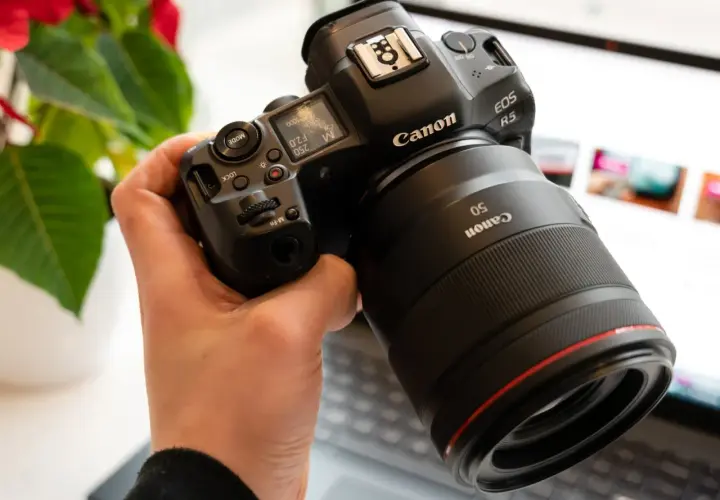 Dłoń trzyma aparat Canon EOS R5 z obiektywem 50mm. W tle widać ekran laptopa.
