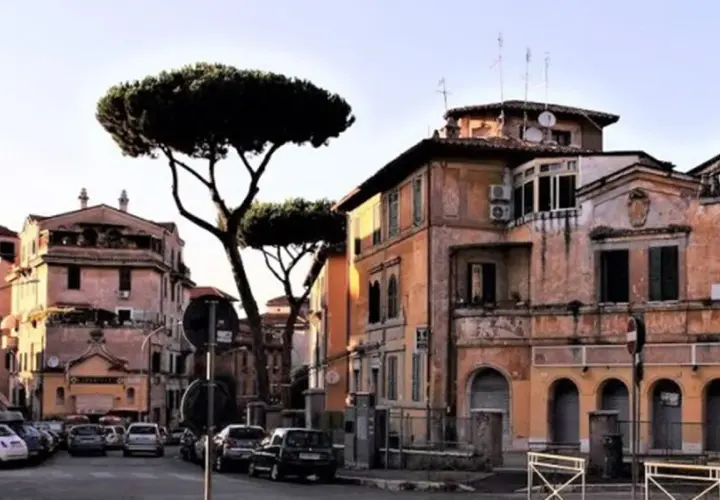 Edifici color ocra e rosa con pini marittimi svettanti, tipici de **la Garbatella**, Roma. Auto parcheggiate lungo la strada.