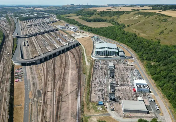Rozległa infrastruktura kolejowa, gdzie pociąg Eurotunelu przygotowuje się do podróży. W tle wzgórza i autostrada.