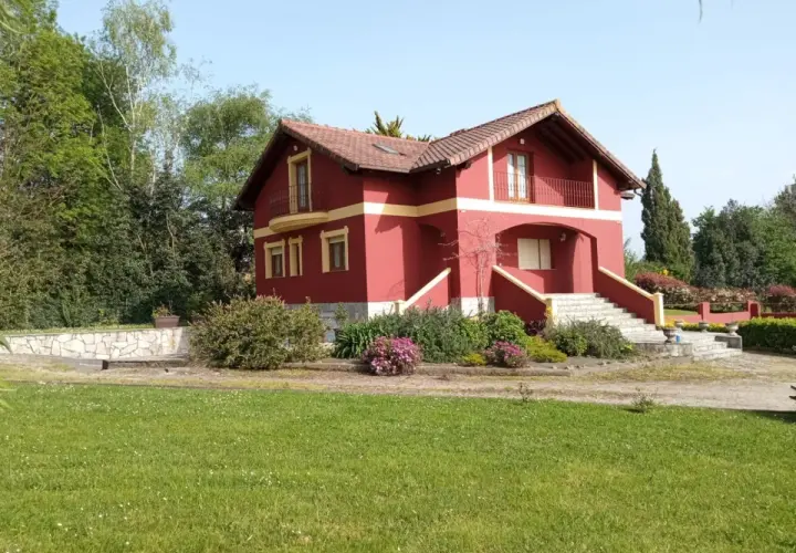 Una casa típica cántabra, de color rojo intenso con detalles amarillos, rodeada de vegetación y un césped verde.