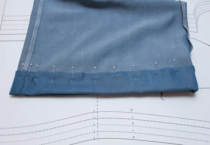 Pins secure the folded hem of blue knit pants, ready for sewing. This image shows a step in how to hem knit pants.