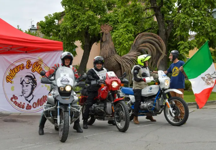 Tre motociclisti pronti per un percorso eroica in moto, con bandiera italiana e un grande gallo di metallo sullo sfondo.