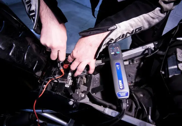 Mani che collegano un caricabatteria Exide alla batteria di una moto. Scopri come caricare batteria moto senza caricabatteria.