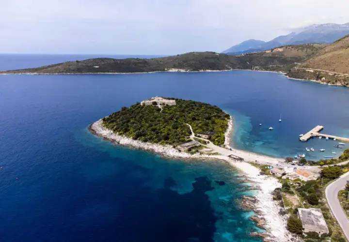 Isla con un castillo en Himare, Albania, rodeada de aguas azules y vegetación exuberante. Un muelle y barcos anclados completan la vista.