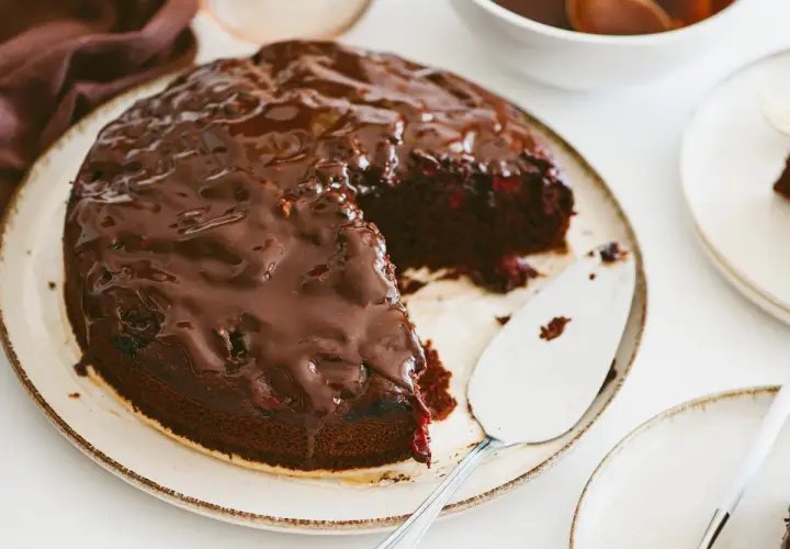 Pyszny brownie z malinami, polane błyszczącą czekoladą, czeka na talerzu. Obok stoi łopatka do ciasta.