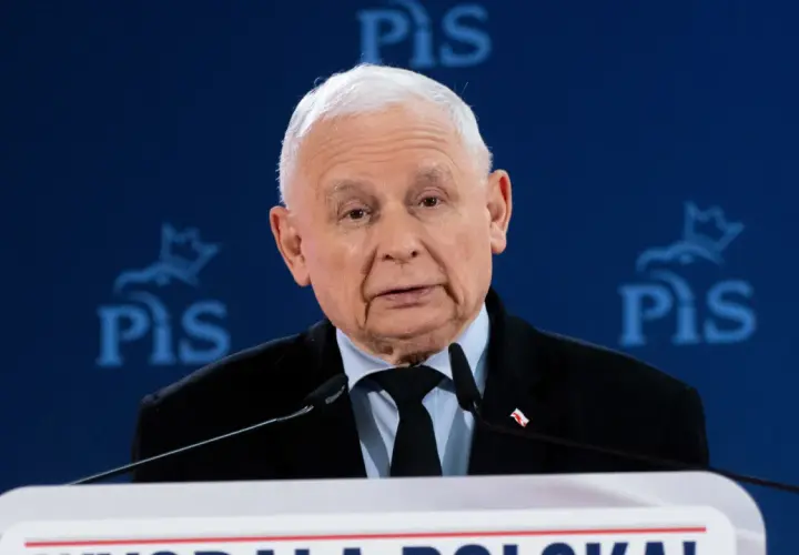 Jarosław Kaczyński w garniturze przemawia za mównicą na tle niebieskiego baneru z logo PiS.
