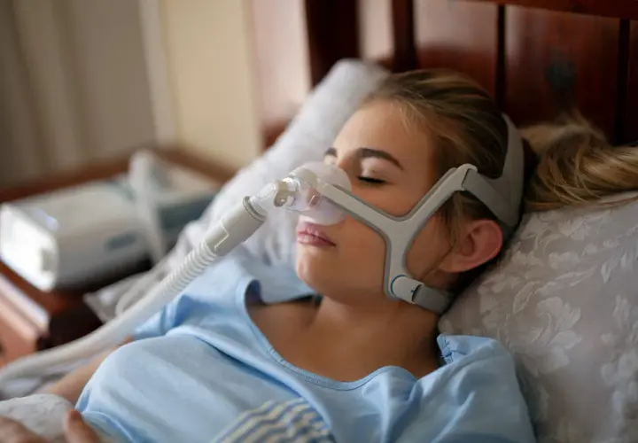 Junge Frau mit Schlafmaske, die gegen Schlafapnoe-Symptome hilft. Ein CPAP-Gerät ist im Hintergrund zu sehen.