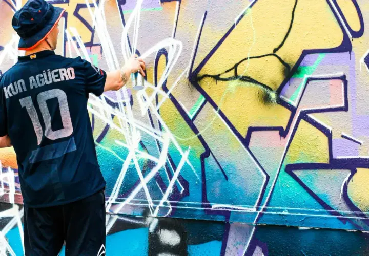 Mann mit Hut und Trikot sprüht mit Spraydose auf bunte Graffiti-Wand.