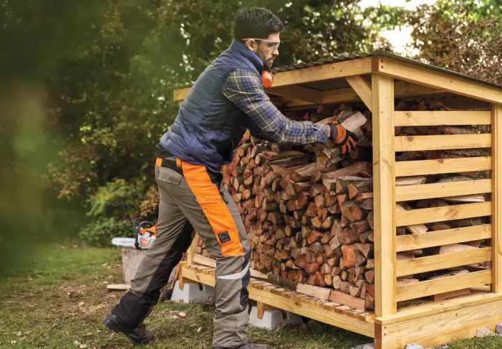 Mann stapelt Holz in einem Holzunterstand. Die Genehmigung für den Holzunterstand ist erteilt, und das Brennholz wird für den Winter vorbereitet.