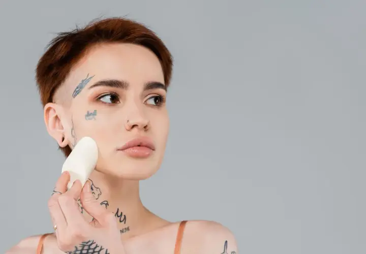 En kvinna med tatueringar applicerar foundation med en svamp för att jämna ut sin underton hud.