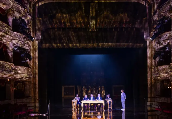 Na scenie teatru, przy złotym stole, siedzi pięć postaci. W tle widać bogato zdobione loże i kurtyny, tworząc atmosferę historycznego przedstawienia.