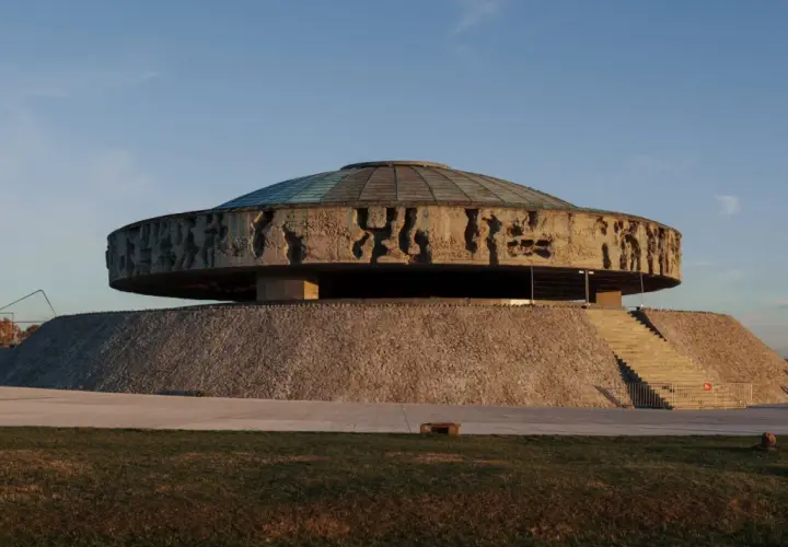 Monumentalne mauzoleum o okrągłej podstawie, z betonowym dyskiem ozdobionym płaskorzeźbami i szklanym dachem.