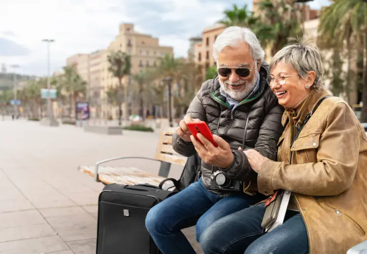 Uśmiechnięta para seniorów sprawdza na telefonie, czy roaming jest domyślnie włączony w Plus, siedząc na ławce z walizką.