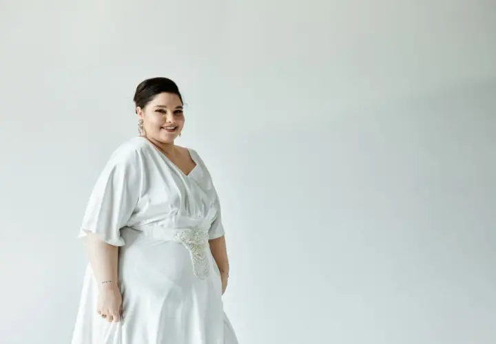 Kobieta w białej sukience z ozdobnym pasem, uśmiecha się do aparatu.