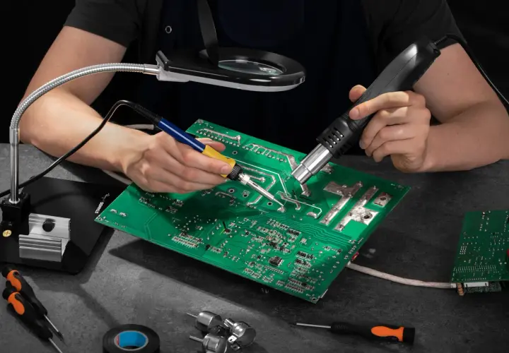 Ręce lutują płytę elektroniczną, używając lutownicy i gorącego powietrza. Widać też lupę, śrubokręty i taśmę izolacyjną – wszystko, co można lutować.