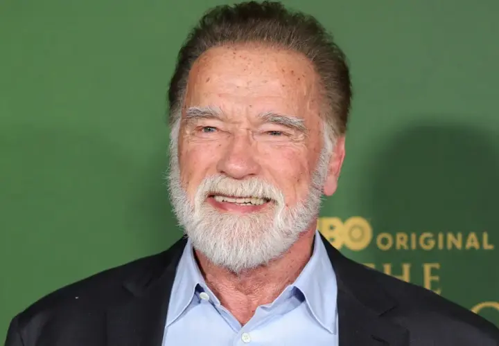 Arnold Schwarzenegger, z siwą brodą i w niebieskiej koszuli, uśmiecha się. W tle zielona ściana z logo HBO.