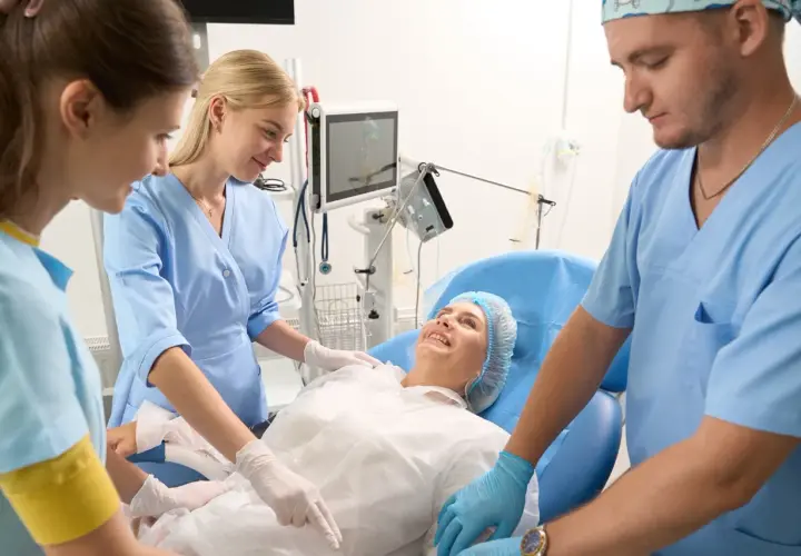 Uśmiechnięta pacjentka w czepku leży na łóżku, otoczona przez zespół medyczny. Opiekun medyczny i dwie pielęgniarki czuwają nad jej komfortem.