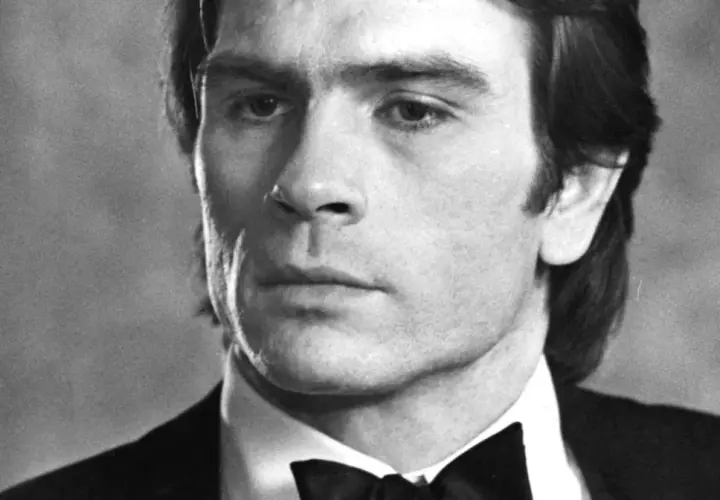 Młody Tommy Lee Jones w czarnym smokingu z muszką, zamyślony.