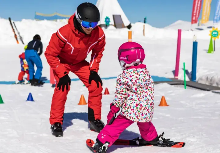 Maestro di sci insegna a bambini a sciare su pista innevata, con coni colorati e bandierine sullo sfondo.