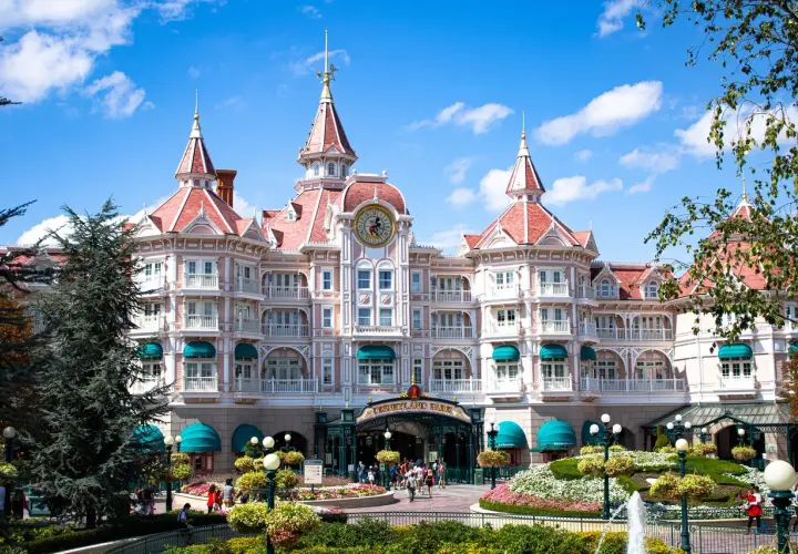 Grand hôtel rose de style victorien avec des tours, un grand cadran d'horloge et des jardins fleuris.